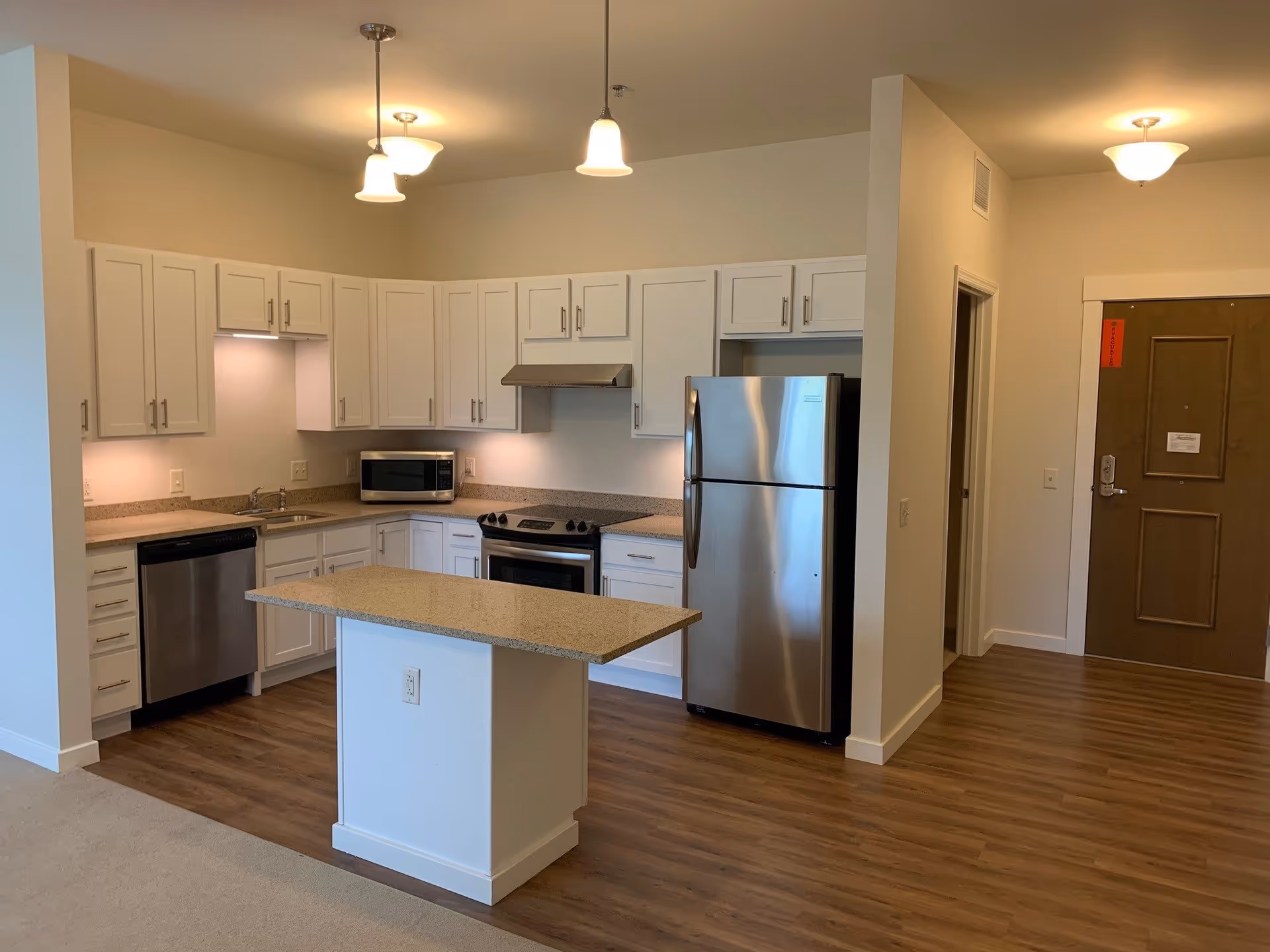 Modern kitchen area with white cabinets, granite countertops, stainless steel refrigerator, dishwasher, stove, and microwave. The kitchen has pendant lights hanging from the ceiling and wood flooring that extends into the adjacent hallway with a brown door.