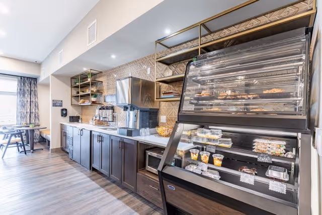 A bright and modern dining area with a long counter featuring a coffee machine, shelves with cups and plates, and a display case containing various pastries and desserts. In the background, there are tables and chairs near a window with patterned curtains.