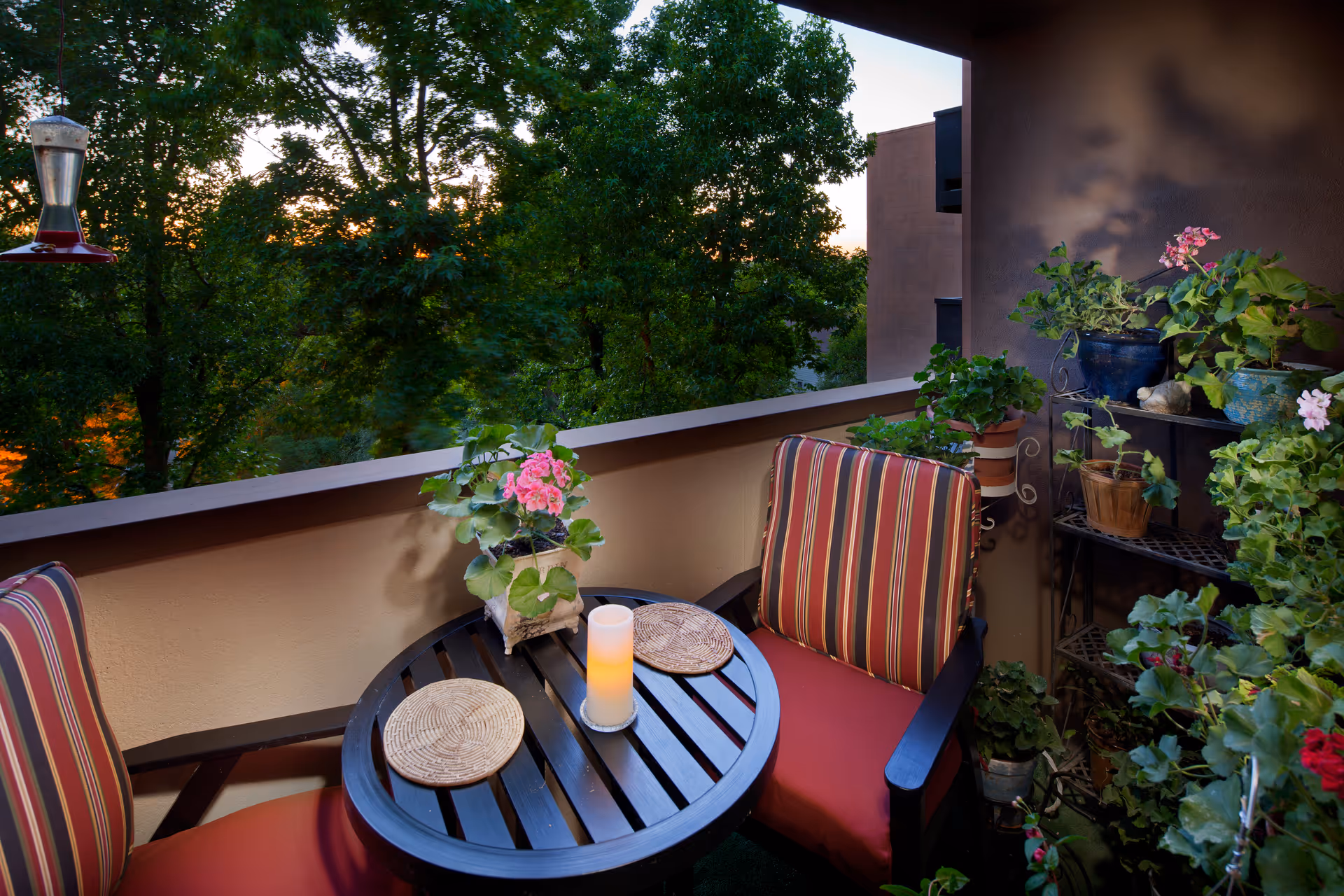 A cozy outdoor balcony area with two striped cushioned chairs and a round black table between them. The table has a lit candle and two woven placemats. Various potted plants and flowers decorate the balcony, with a green leafy view of trees in the background during sunset.