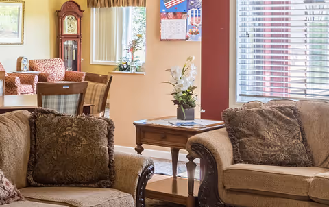 A cozy senior living common area featuring two beige upholstered armchairs with patterned cushions, a wooden side table with a floral arrangement, a grandfather clock, and a dining table with chairs in the background. The room has large windows with blinds and a wall calendar with patriotic decorations.