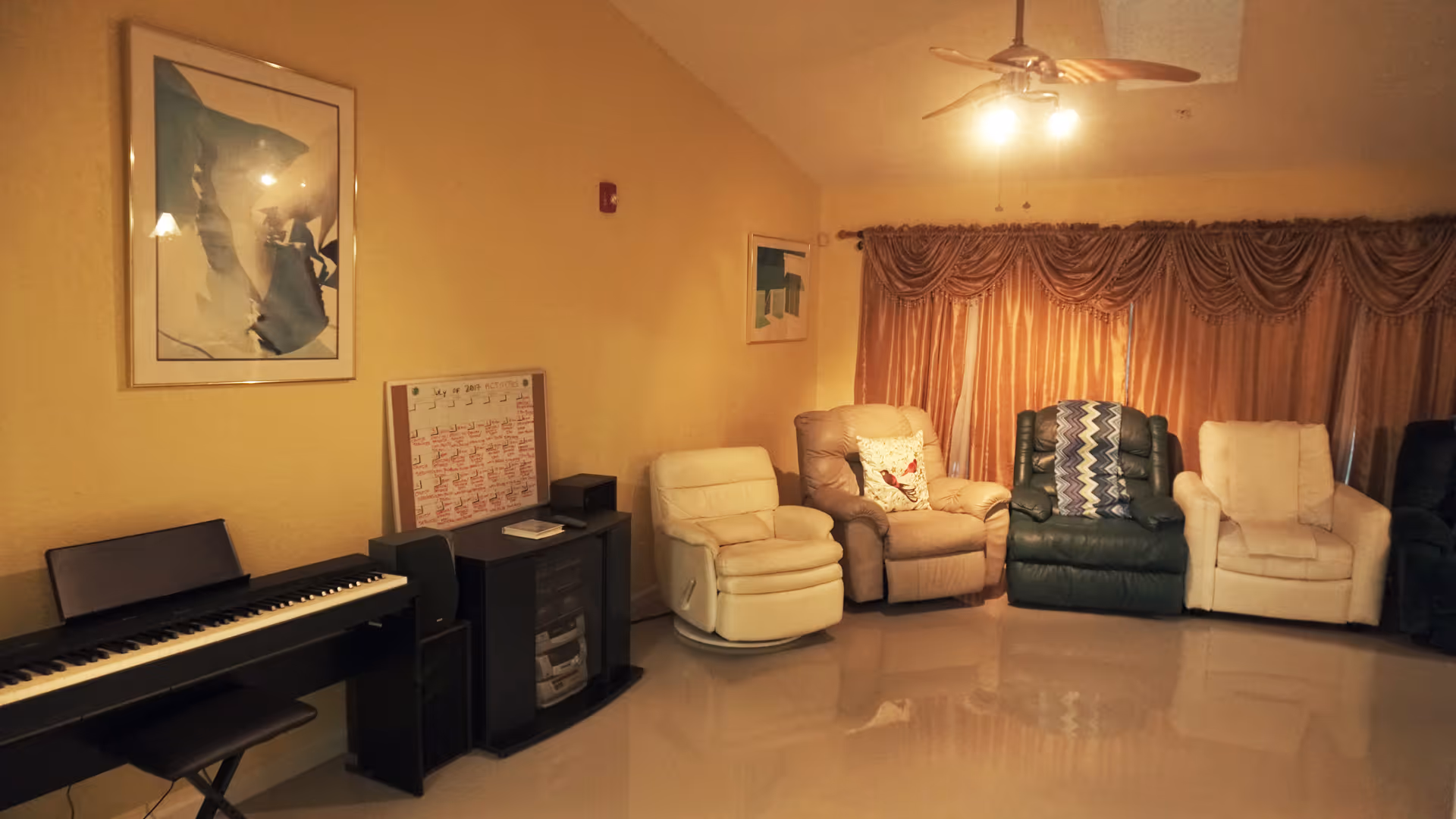 A cozy living room with five recliner chairs arranged in a row against a wall with large, heavy curtains. There is a ceiling fan with a light fixture above. On the left side, there is an electronic keyboard on a stand with a bench, a small cabinet with speakers, and a whiteboard calendar on the wall. The walls are painted a warm beige color.