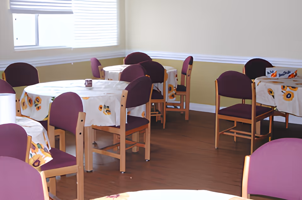 A dining room with several round tables covered with floral tablecloths. Each table is surrounded by wooden chairs with purple cushions. There is a window with blinds letting in natural light, and the walls are painted in a light beige color with white trim.
