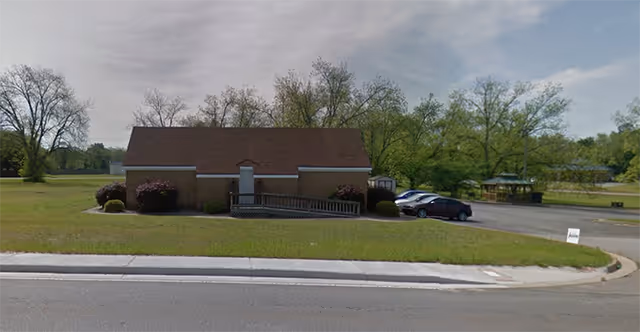 Single-story brick building with a brown shingled roof, a central white door with a wheelchair ramp, surrounded by grass and trees, with a parking lot and a gazebo visible to the right.