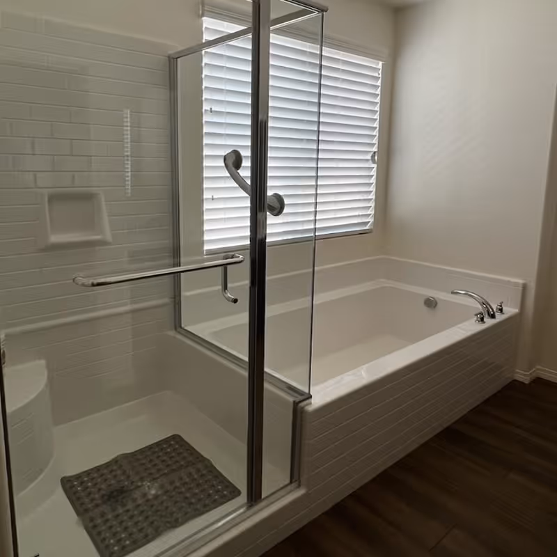 A clean bathroom featuring a glass-enclosed shower with a grab bar and a built-in seat, adjacent to a white bathtub with silver fixtures. A window with white blinds is above the bathtub, and the floor has dark wood-like flooring.