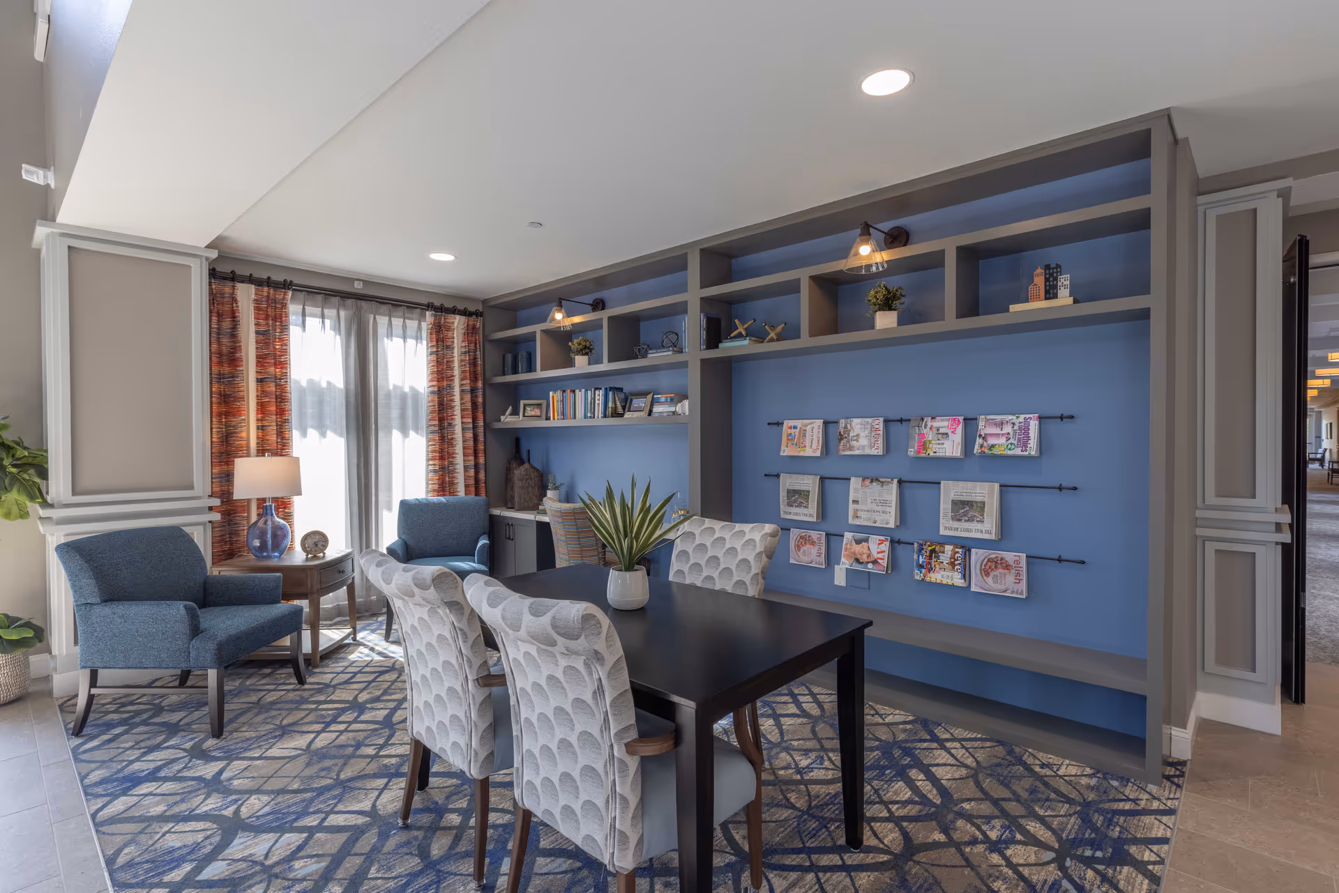 A bright communal seating area with a table and patterned chairs, two armchairs by curtains, and built-in blue shelving displaying books and magazines.