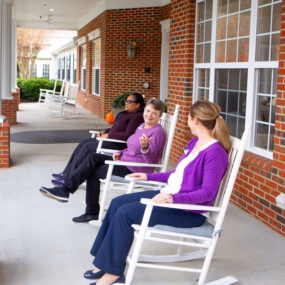 Three women sitting and chatting on white rocking chairs on a covered porch with brick walls and large windows at Spring Arbor of Wilson.