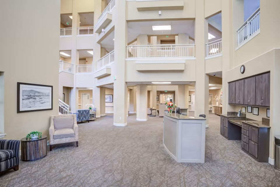 Spacious and well-lit interior lobby area of a senior living facility with beige walls, carpeted floors, a reception desk with flowers, seating areas with chairs and tables, and multiple floors visible with white railings.