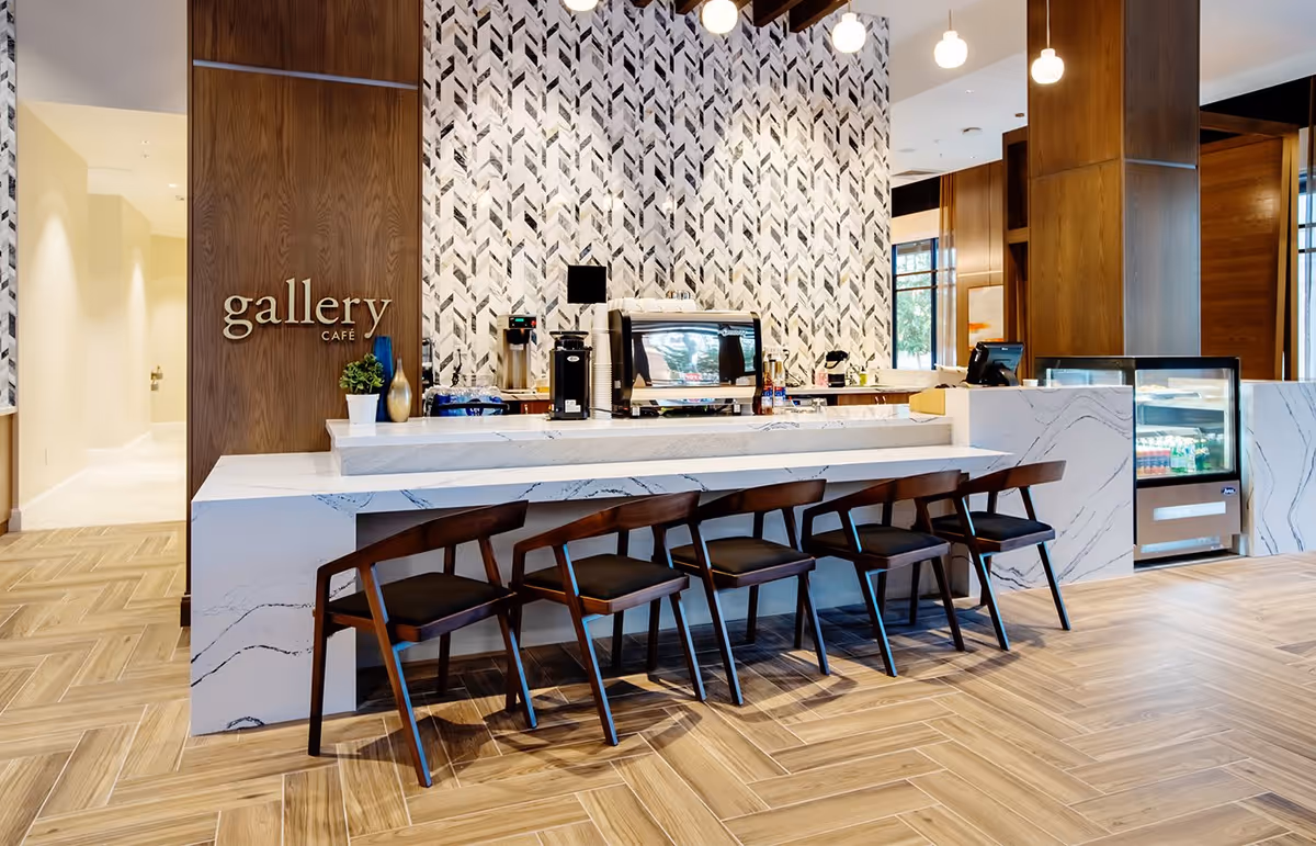 Modern cafe area with a marble counter and five wooden chairs with black cushions. Behind the counter is a coffee machine, cups, and other cafe equipment. The wall behind features a geometric patterned tile design, and a wooden panel with the words 'gallery cafe' in gold letters. The floor has a light wood herringbone pattern.