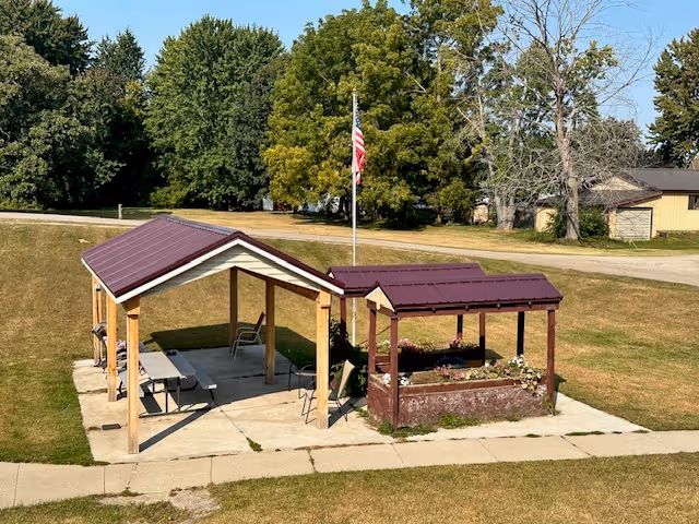 Outdoor pavilion with two connected covered structures featuring maroon roofs and wooden supports, one with picnic tables and chairs underneath, and the other with a raised flower bed. An American flag on a flagpole is visible behind the pavilion, surrounded by grass and trees in the background.