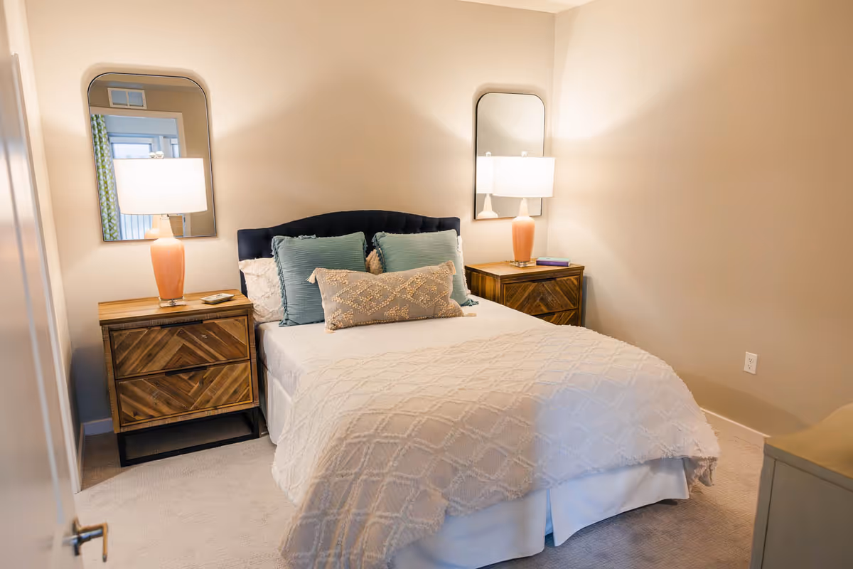 A neatly made bedroom with a bed, decorative pillows, two wooden nightstands with lamps, and wall mirrors on beige walls.
