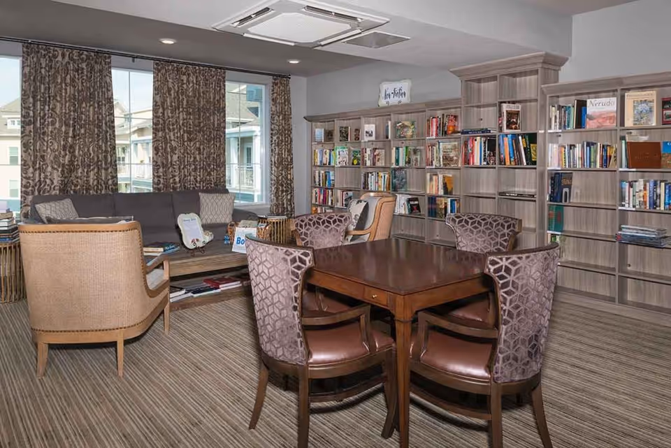 A cozy reading and sitting area in a retirement community featuring a wooden table with four upholstered chairs, a sofa with cushions, two armchairs, and multiple bookshelves filled with books. Large windows with patterned curtains allow natural light into the room.