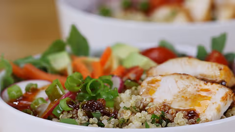 Close-up of a bowl of quinoa salad topped with grilled chicken, chopped green onions, cherry tomatoes, avocado slices, and shredded carrots.