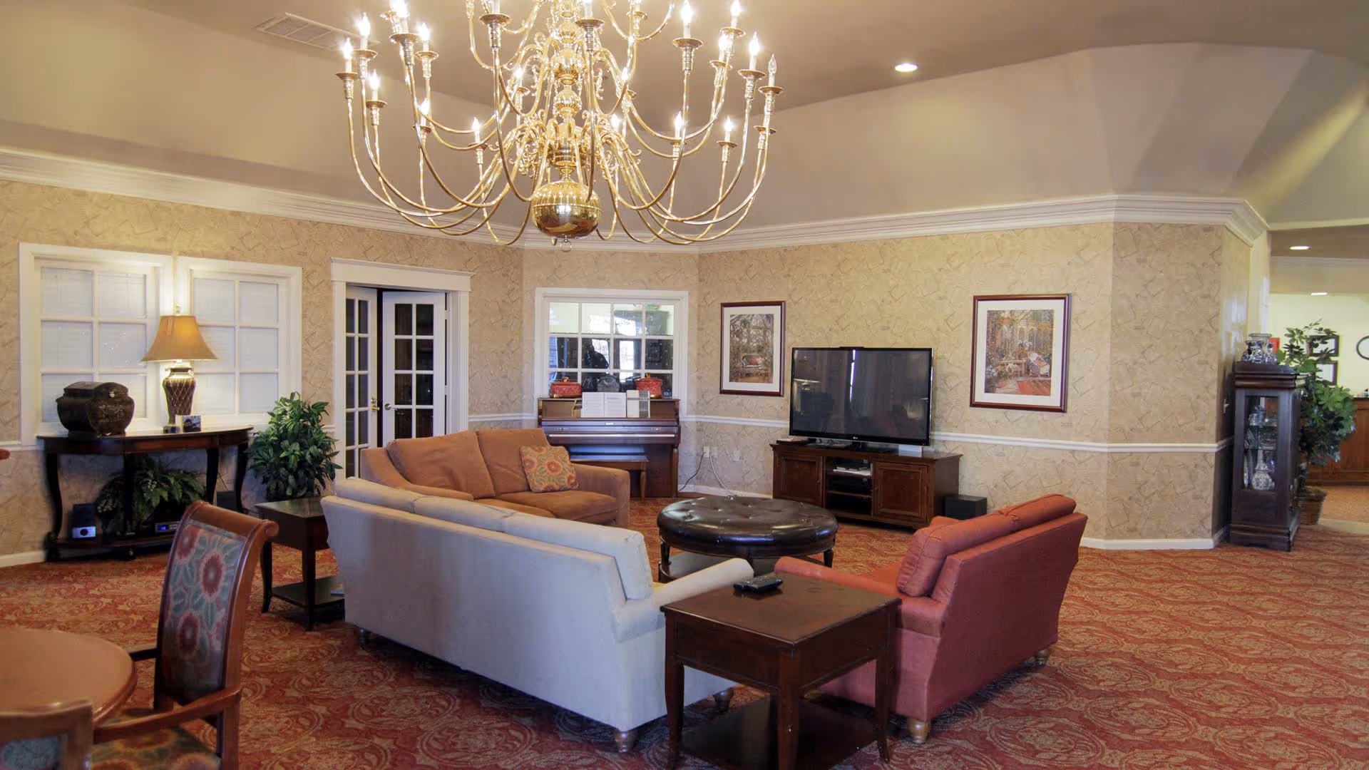 A senior living facility common area with a large chandelier hanging from the ceiling, several upholstered sofas and chairs arranged around a round ottoman, a flat-screen TV on a wooden stand, a piano against the wall, framed artwork, and decorative plants.