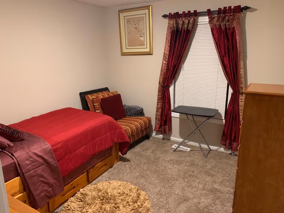 A cozy bedroom with a single bed covered in red bedding, a striped armchair with red and brown cushions, a small folding table in front of a window with red and gold curtains, a framed picture on the wall, and a wooden dresser. The floor is carpeted and there is a round beige rug near the bed.