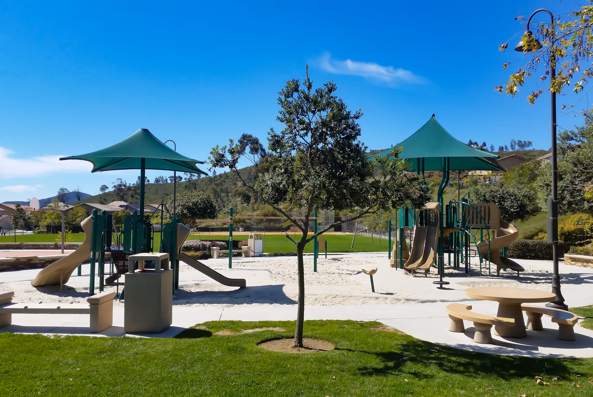 Outdoor playground area with slides, climbing structures, and shaded canopies surrounded by green grass, trees, and hills under a clear blue sky.