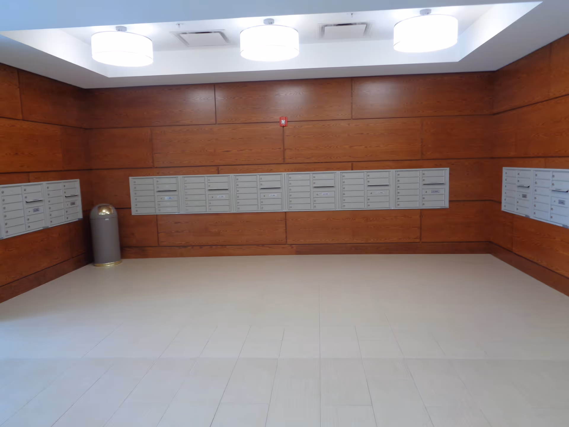 A clean interior mailroom with rows of wall-mounted mailboxes, wood-paneled walls, a trash bin, tiled floor, and ceiling lights.