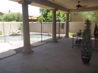Covered outdoor patio overlooking a pool, with columns, railing, and potted plants.