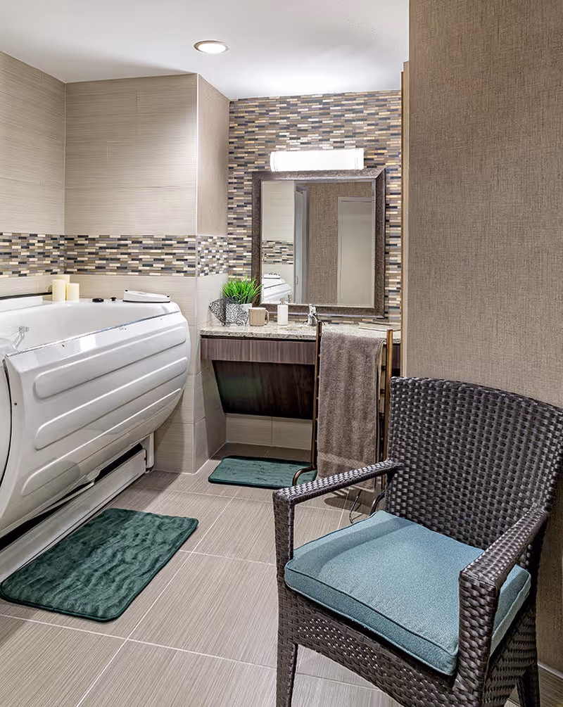 Modern bathroom with a walk-in tub, vanity and mirror, tiled walls, and a wicker chair with cushions.