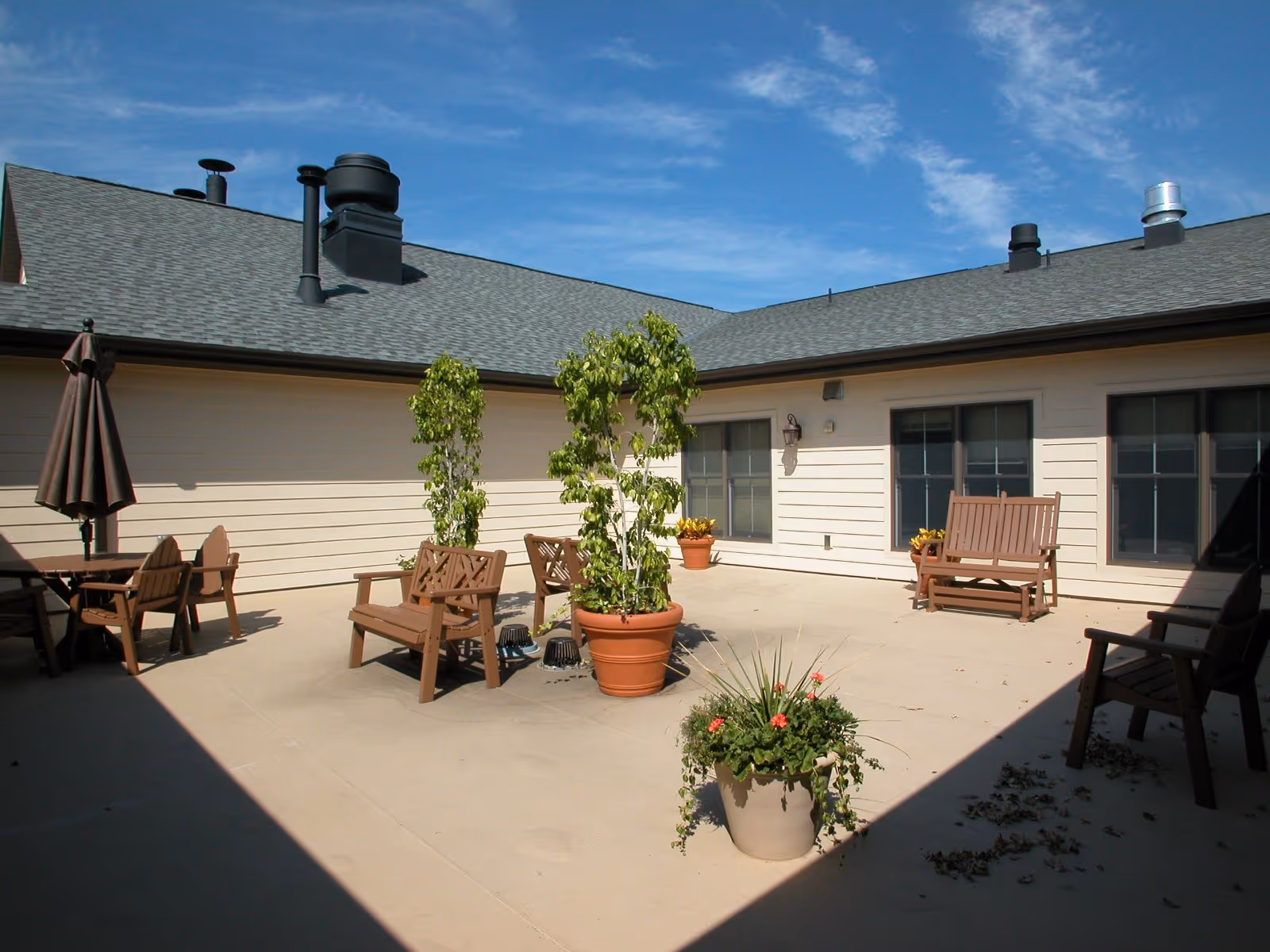 Outdoor patio area with several wooden benches and chairs, potted plants, and a closed umbrella under a clear blue sky. The patio is surrounded by a building with beige siding and multiple windows.