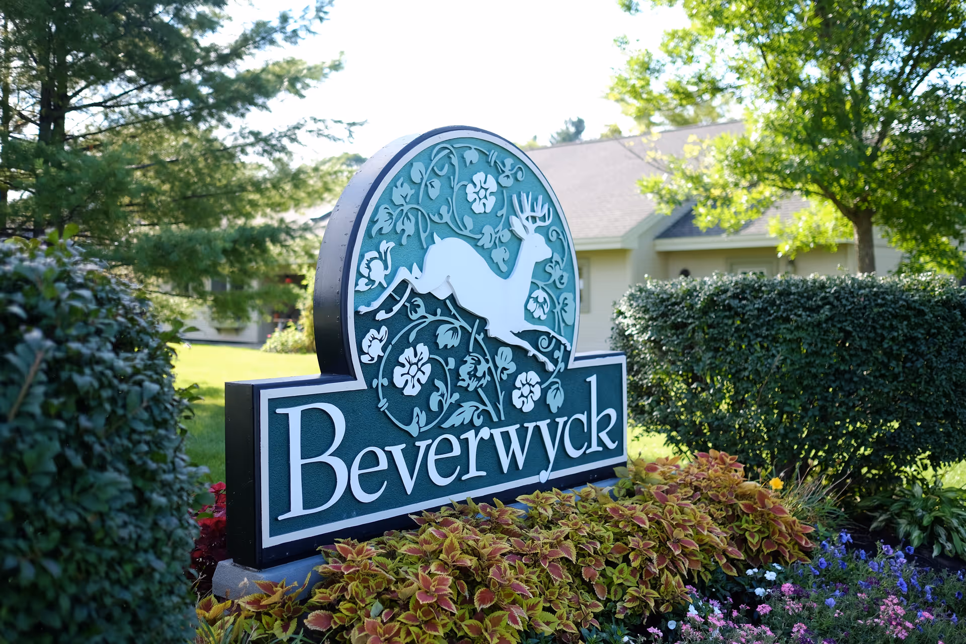 A decorative outdoor sign for Beverwyck featuring a white deer silhouette surrounded by floral patterns, set among green bushes and colorful flowers with a building and trees in the background.