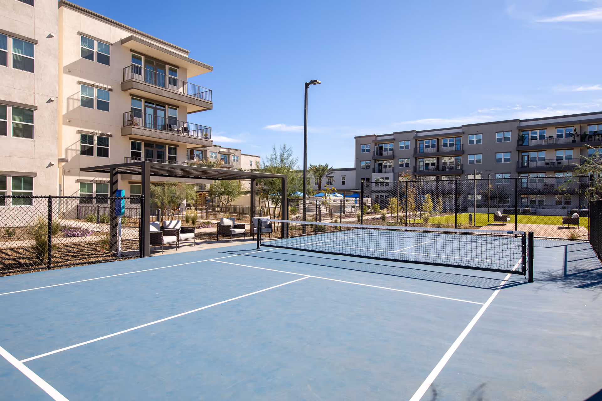 Outdoor fenced tennis/pickleball court with seating and a pergola in front of multi-story apartment buildings under a clear sky.