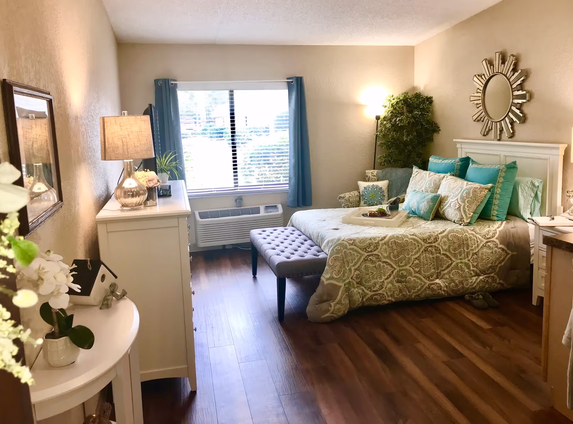 Bright furnished bedroom with a bed dressed in patterned bedding and teal pillows, a bench, dresser and window with curtains.
