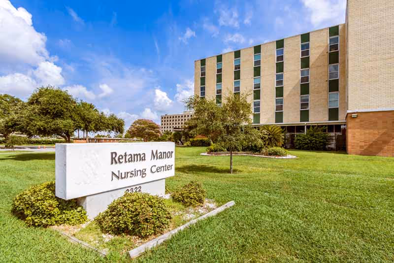 Exterior view of Retama Manor Nursing Center building with a large sign in the foreground on a grassy lawn. The building is multi-story with many windows and surrounded by trees and landscaping under a partly cloudy blue sky.