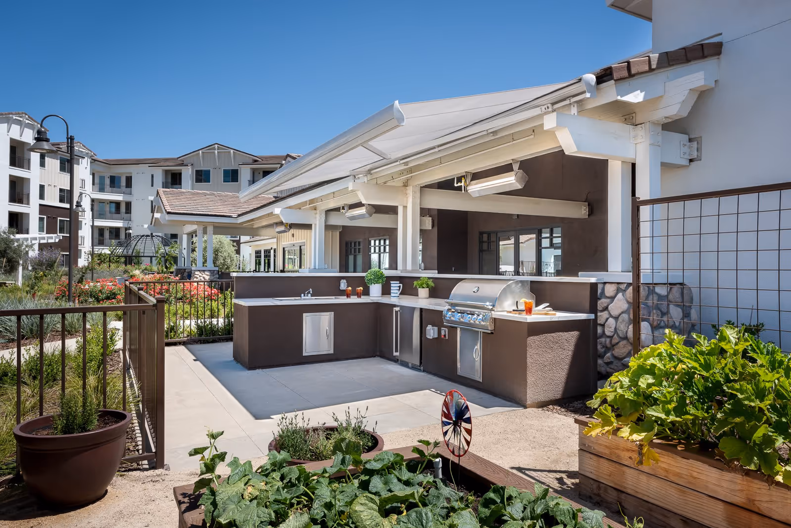 Outdoor kitchen area with a built-in grill, sink, and countertop under a white pergola. Surrounding the kitchen are garden beds with green plants and a clear blue sky overhead. In the background, there are multi-story residential buildings and landscaped pathways.