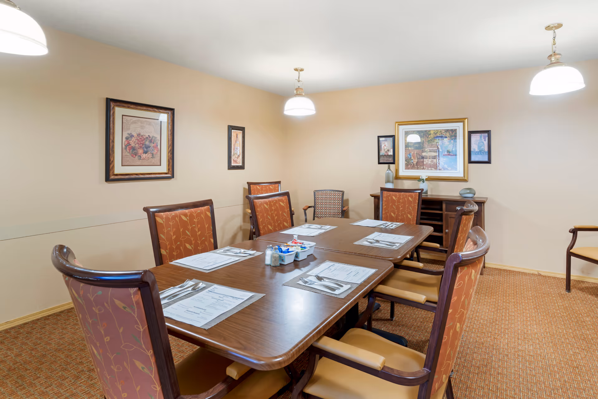 A dining room with a rectangular wooden table set for six people with placemats, menus, and silverware. The chairs have wooden frames with orange patterned upholstery. The room has beige walls adorned with framed artwork and two hanging pendant lights above the table. There is a wooden sideboard against the far wall.