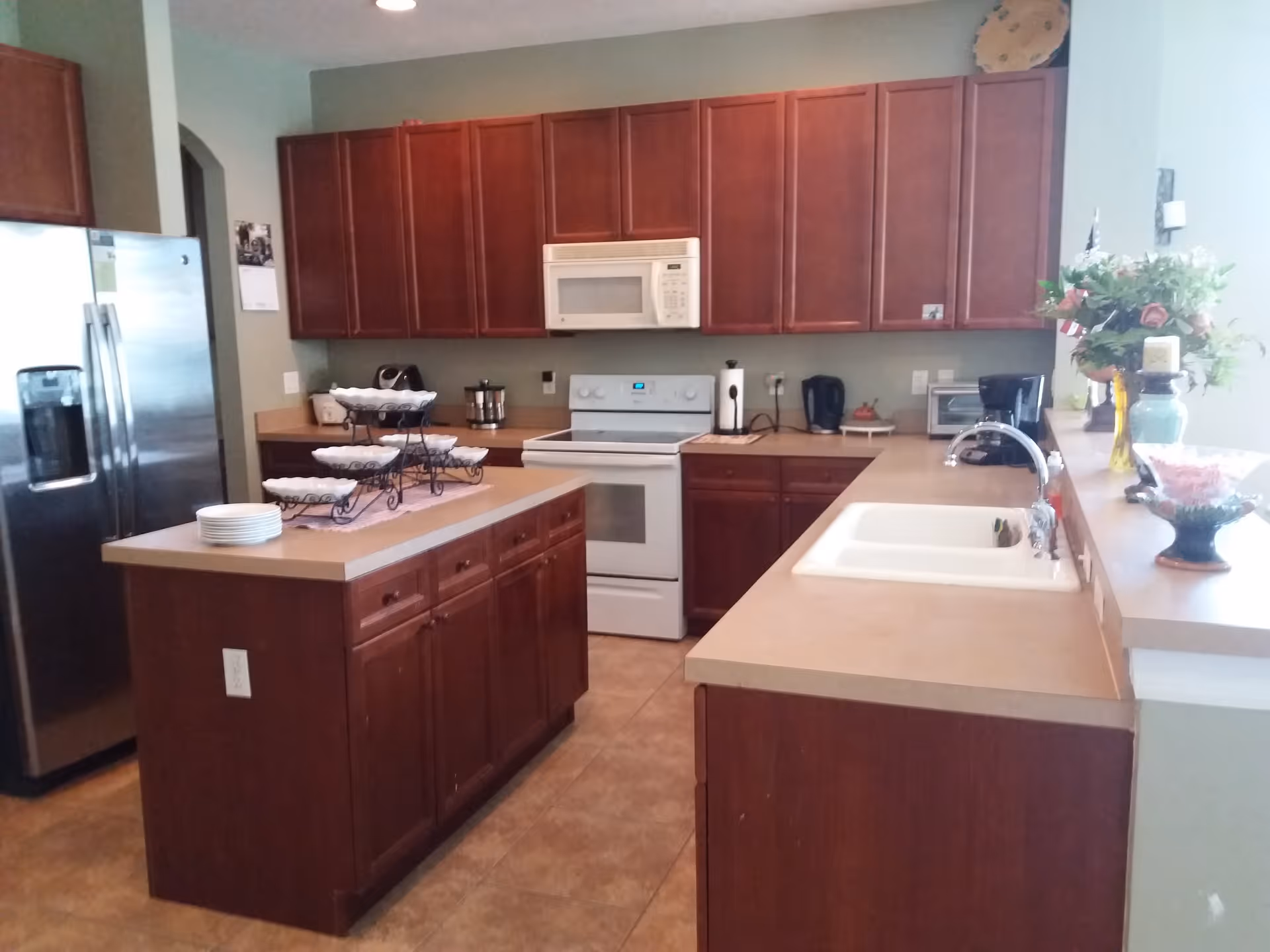 A kitchen with wooden cabinets, a white stove and microwave, a stainless steel refrigerator, a kitchen island with plates and a tiered serving tray, a double sink, and various small appliances and decorative items on the countertops.