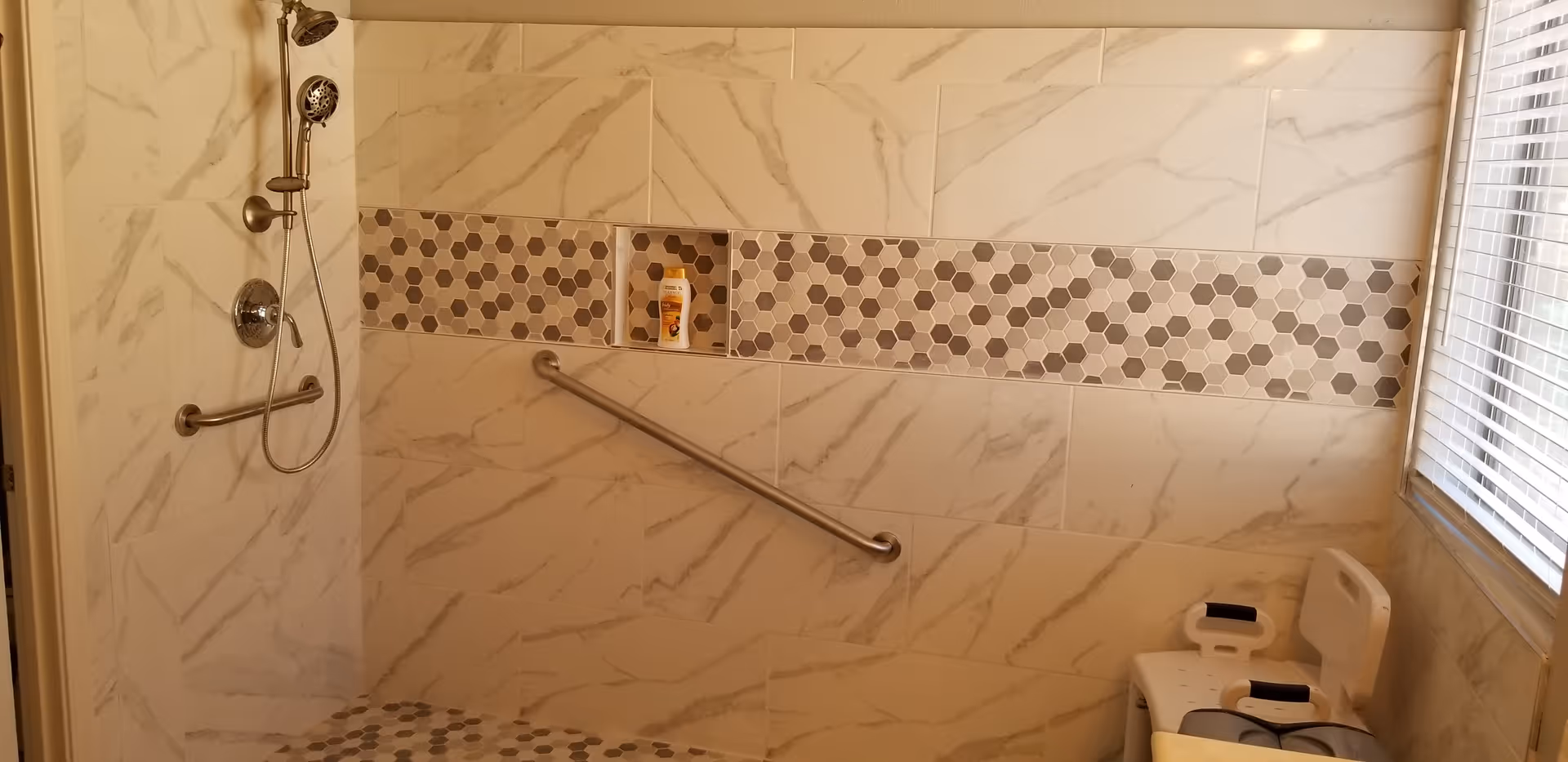 A bathroom shower area with marble-patterned tiles and a horizontal strip of hexagonal mosaic tiles. There is a handheld showerhead, a grab bar on the wall, a built-in niche holding a bottle, and a shower chair near a window with blinds.
