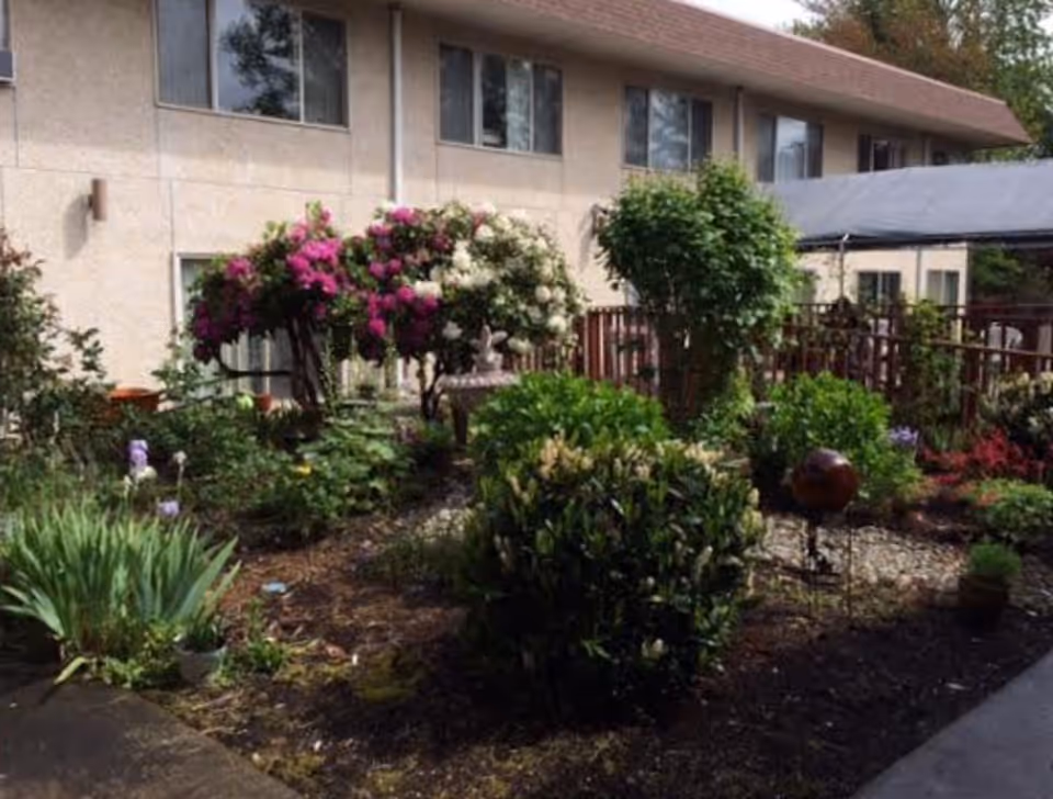 A garden area with various green shrubs, flowering plants including pink and white flowers, and a small birdbath or fountain in the center. The garden is adjacent to a beige two-story building with several windows. There is a wooden fence and a covered walkway on the right side.