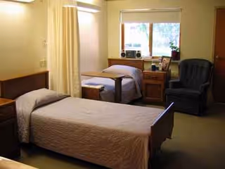 Two single beds with a privacy curtain, dresser, and chair in a shared elder care bedroom.