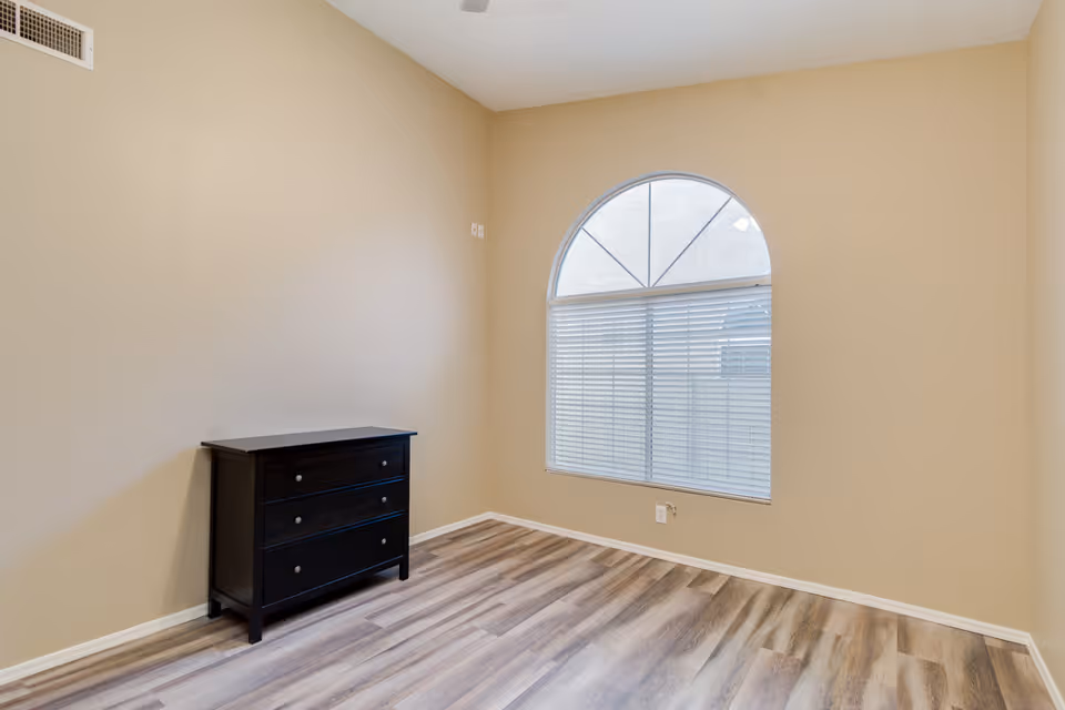 Empty room with beige walls, a large arched window with blinds, wood-patterned flooring, and a small black three-drawer dresser against one wall.