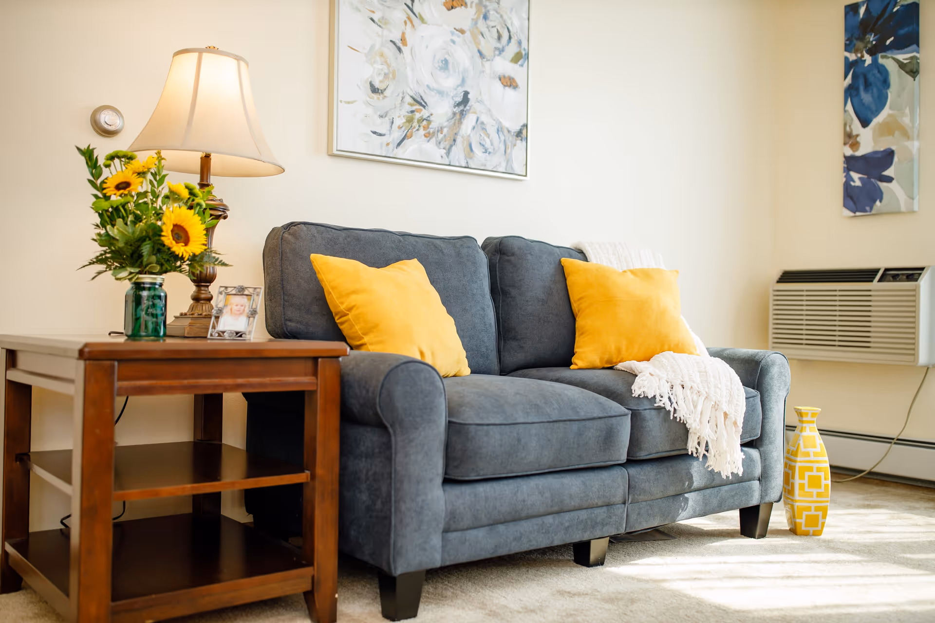 A cozy living room area featuring a dark gray sofa with two yellow throw pillows and a white knitted blanket draped over one arm. Next to the sofa is a wooden side table with a lamp, a vase of sunflowers, and a framed photo. The wall behind has floral artwork, and there is a yellow decorative vase on the floor near a wall-mounted air conditioning unit.