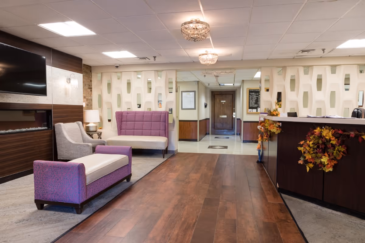 A reception area in a senior living facility with a wooden floor, a dark wood reception desk decorated with autumn leaf garlands, purple and beige upholstered seating, a wall-mounted TV above a modern fireplace, and decorative ceiling lights.