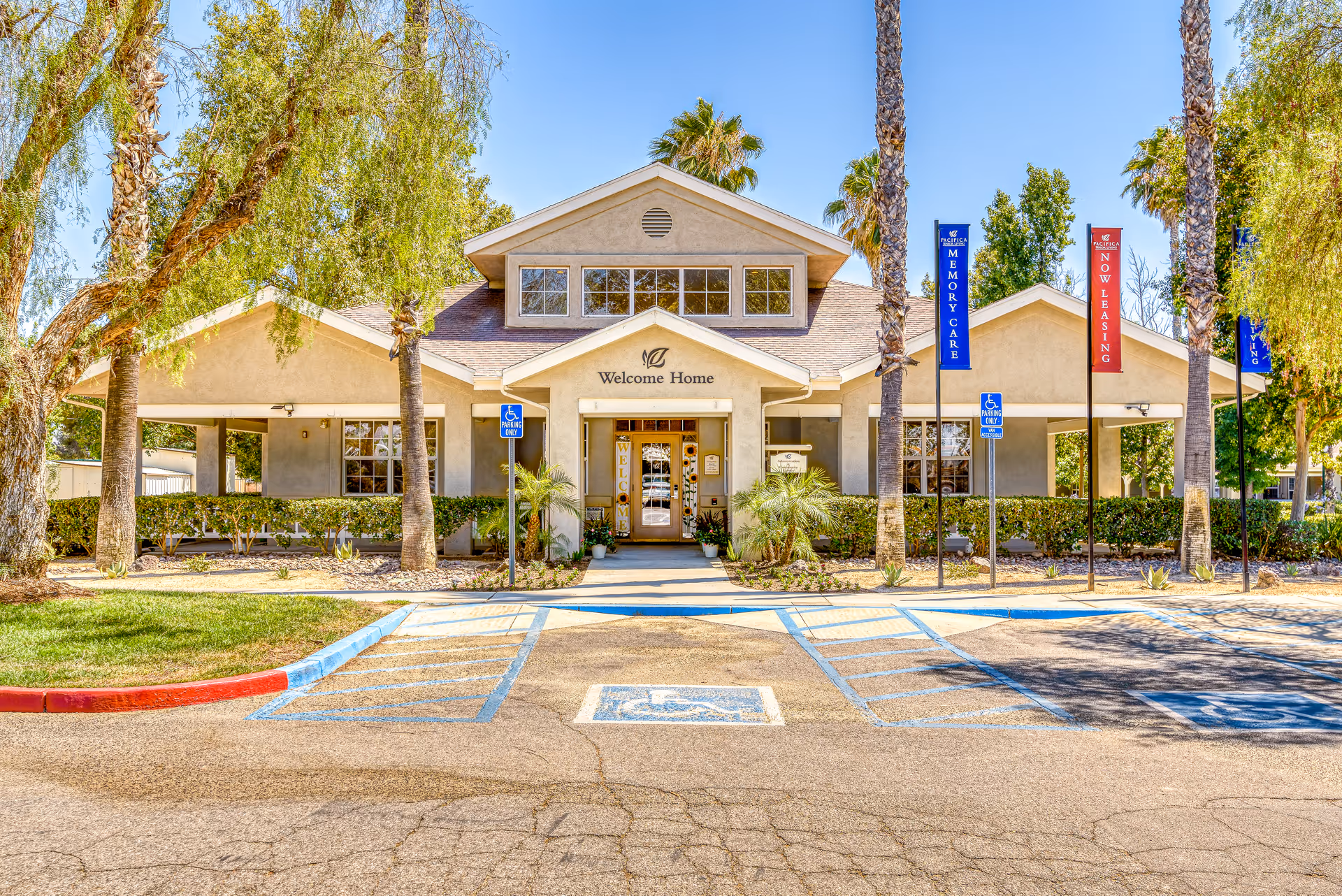Front entrance of a single-story senior living building with a 'Welcome Home' sign, palm trees, and marked handicapped parking spaces.