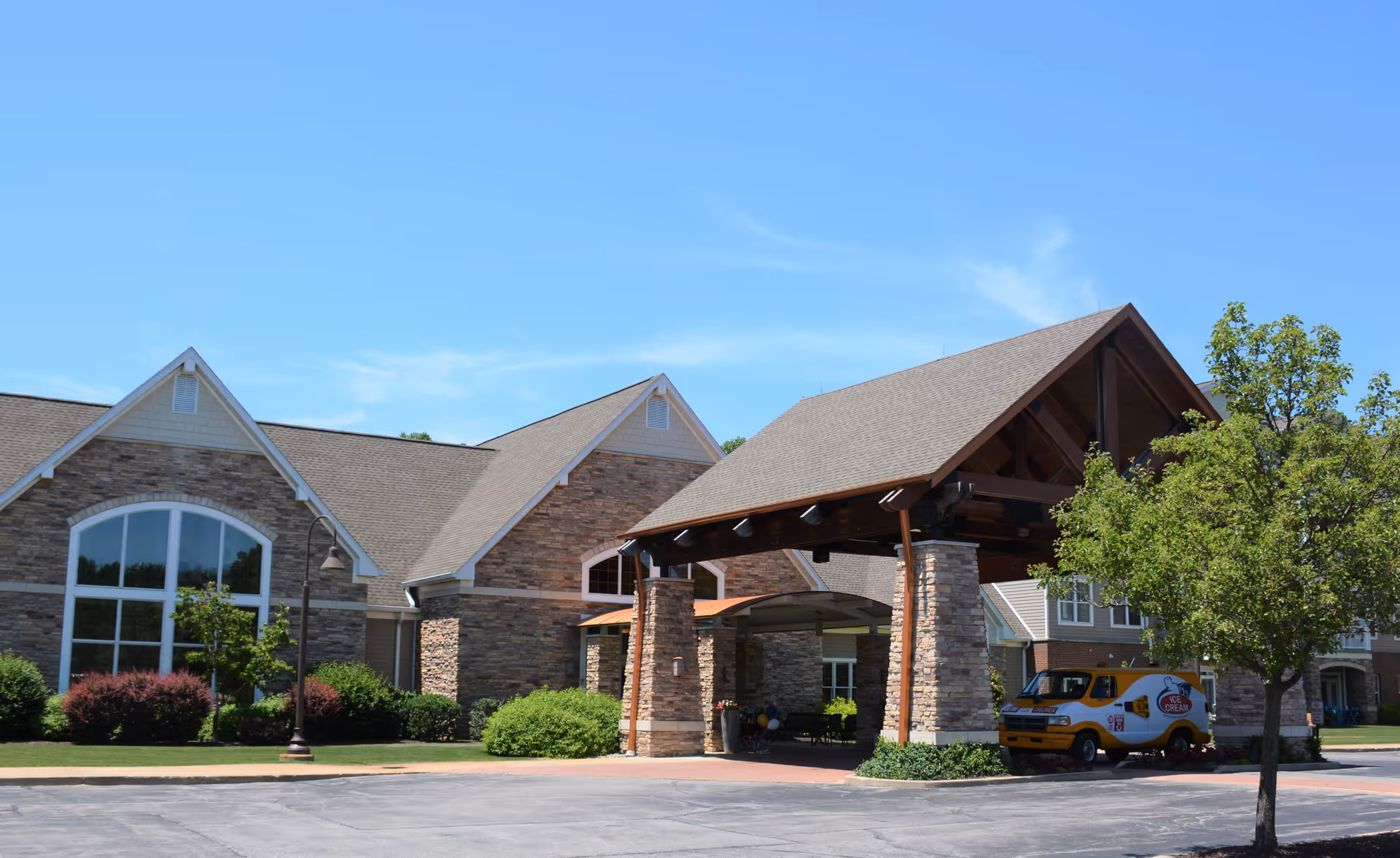 Exterior view of a senior living facility with stone and brick architecture, featuring a covered entrance supported by stone pillars. There is a tree and bushes in the landscaped area, and a yellow ice cream truck is parked near the entrance under the covered area. The sky is clear and blue.