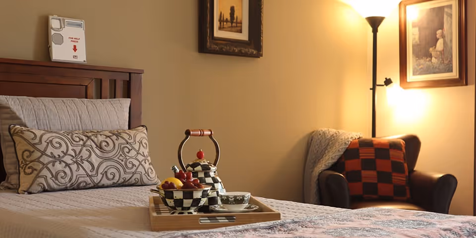 A cozy bedroom in an assisted living facility featuring a neatly made bed with decorative pillows and a tray holding a teapot, bowl of fruit, and a cup. In the background, there is a comfortable armchair with a checkered pillow and a blanket draped over it, a floor lamp, and framed artwork on the wall.
