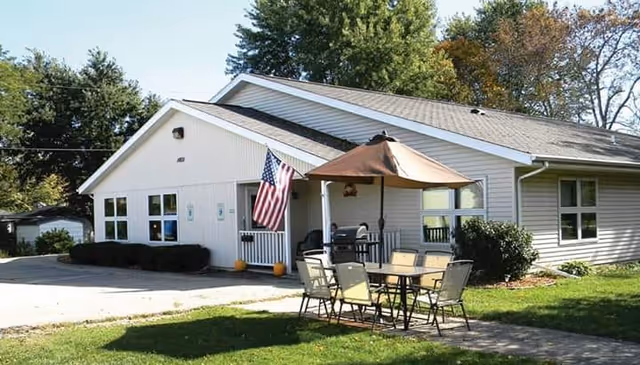 Single-story light-colored building with a sloped roof, an American flag near the entrance, and an outdoor seating area with a table, chairs, and a large umbrella on a grassy lawn.