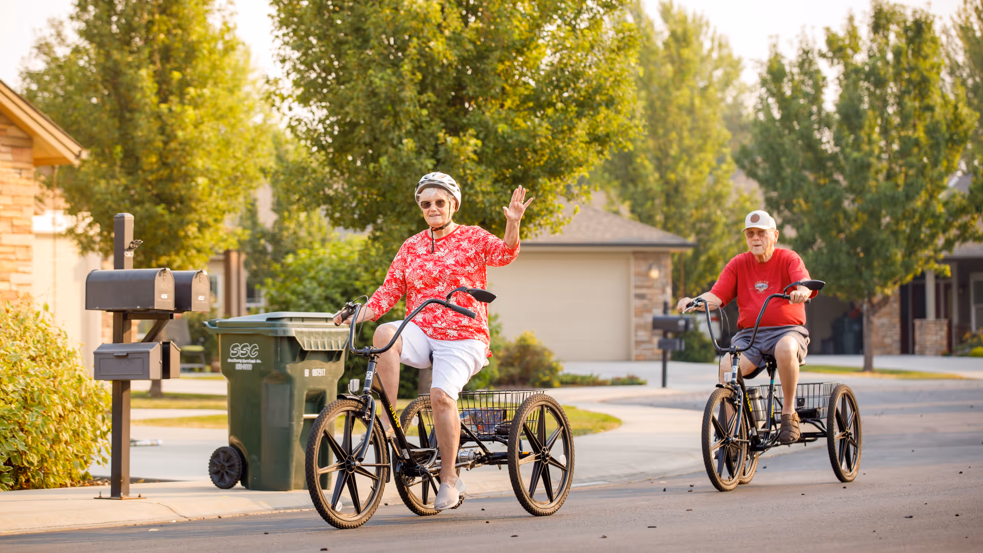 Two elderly individuals riding three-wheeled bicycles on a residential street lined with trees and houses. One person is waving and wearing a helmet and sunglasses, while the other is wearing a cap. The scene is bright and sunny with green foliage in the background.