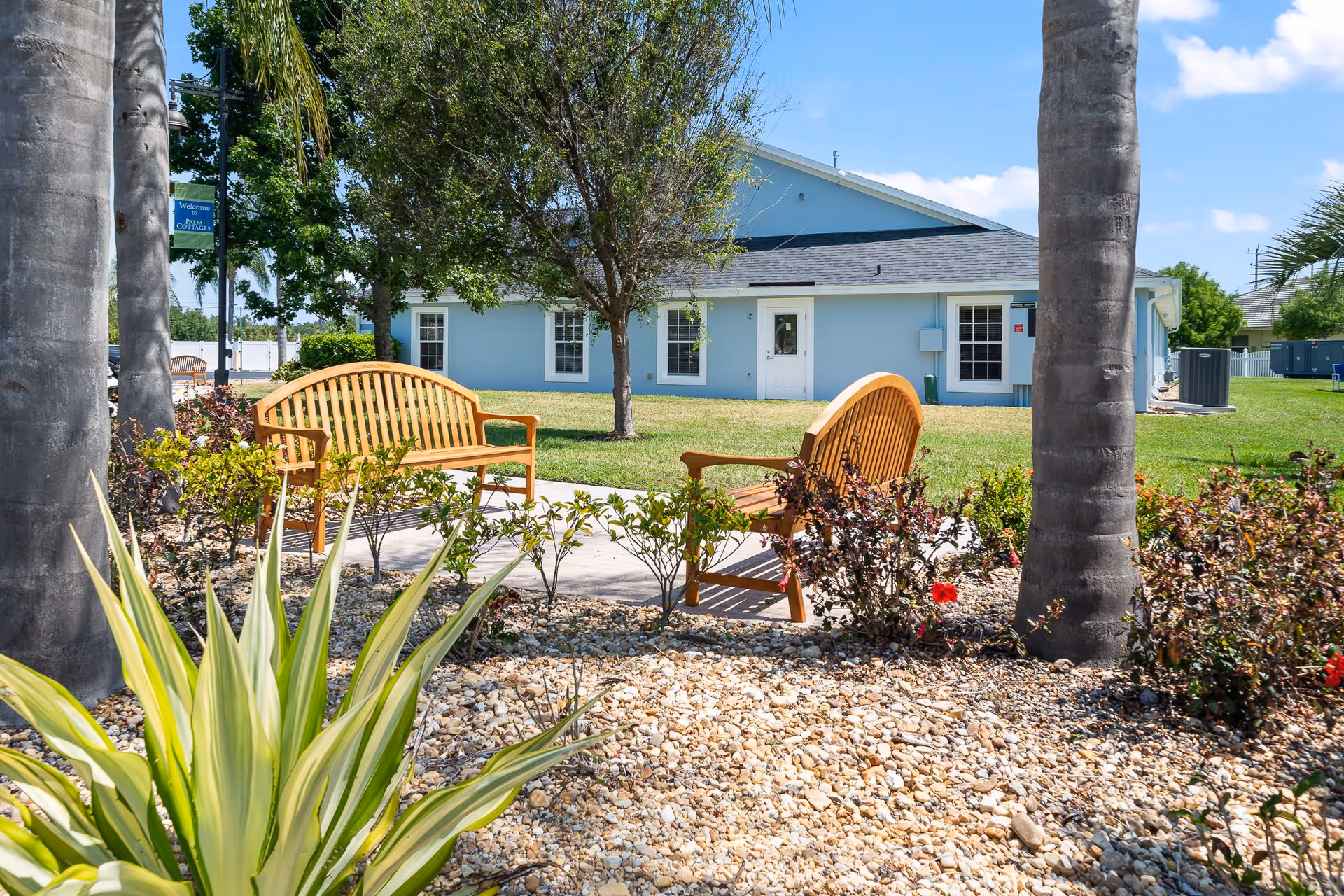 Outdoor seating area with two wooden benches on a concrete pad surrounded by small bushes and plants, with a light blue building and trees in the background under a partly cloudy sky.