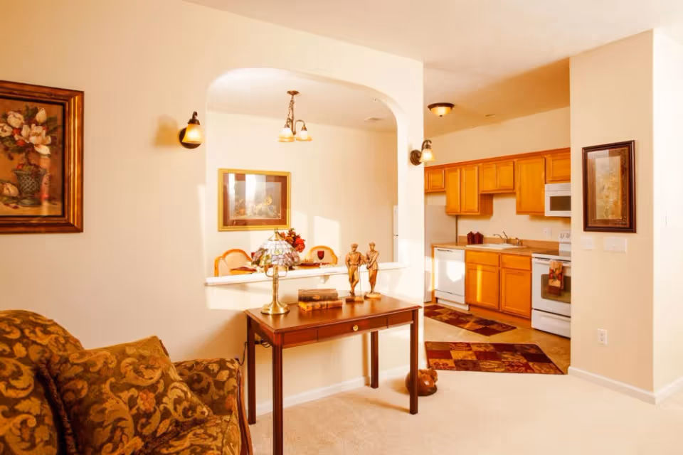 Interior view of a senior living facility showing a cozy living room area with a patterned sofa and a wooden table with decorative items. The room opens into a kitchen with wooden cabinets, white appliances, and a tiled floor with patterned rugs. The walls are decorated with framed artwork and the space is warmly lit.