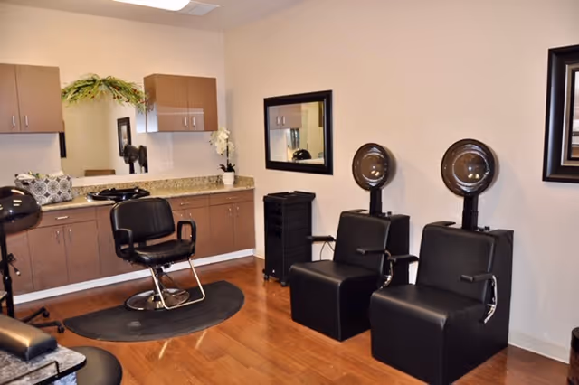 Interior of a salon area in a senior living facility with two black hair drying chairs and one styling chair in front of a counter with cabinets and a large mirror. The floor is wooden, and the walls are light-colored with a framed mirror and a small plant on the counter.