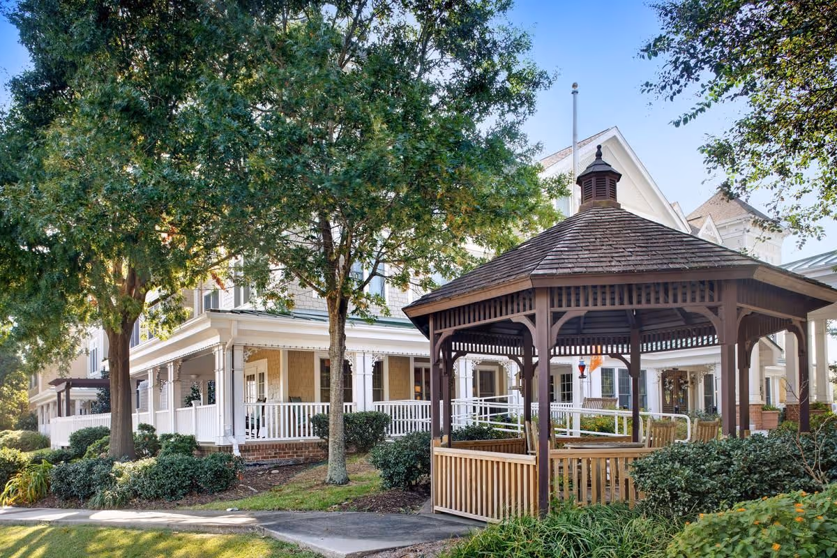 Front exterior of a senior living building with a wooden gazebo, wraparound porch, trees, and landscaping.