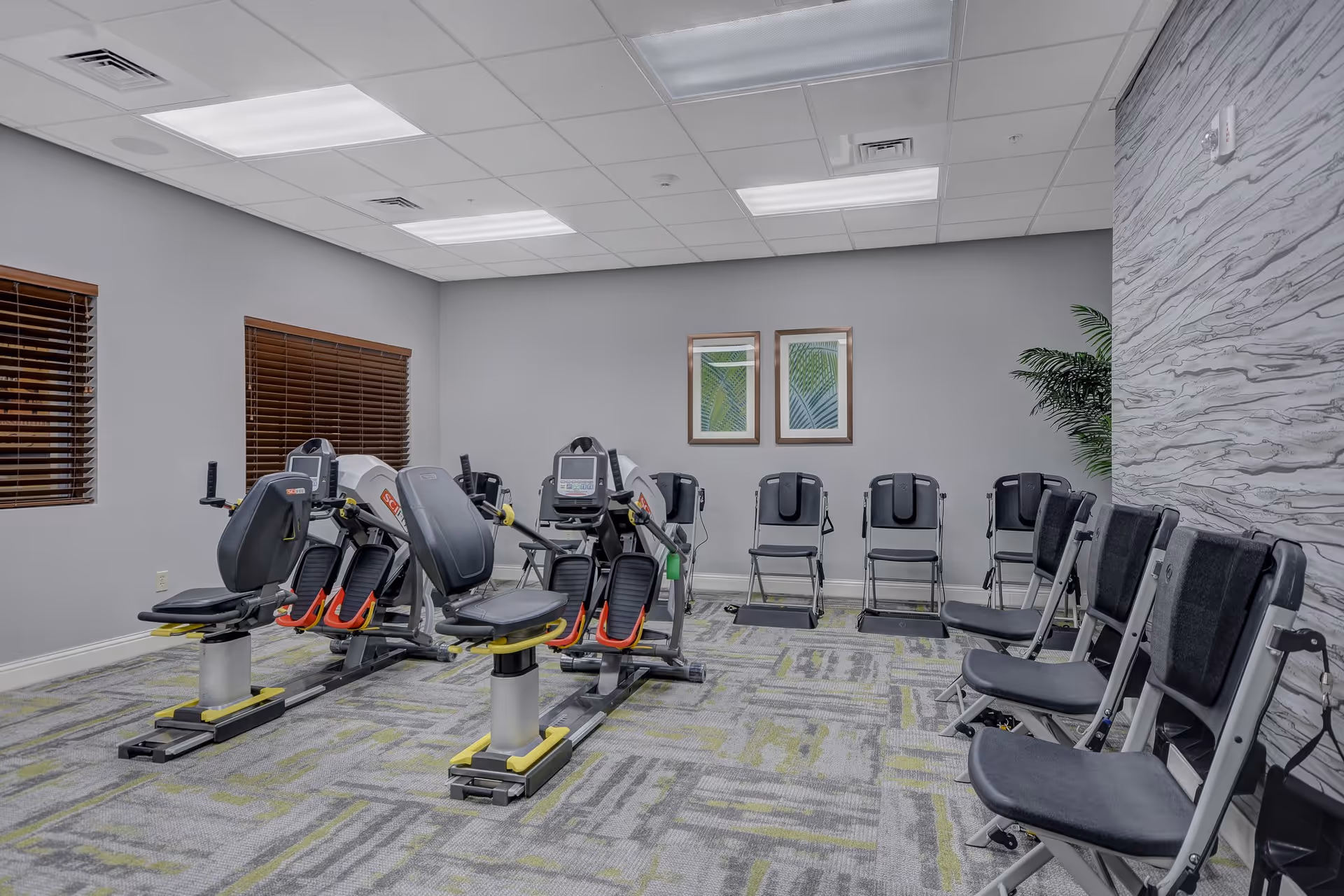 A fitness room with two recumbent exercise bikes in the center and several black folding chairs arranged along the walls. The room has gray walls, two framed pictures of green leaves, a patterned carpet with gray and green tones, and wooden blinds on the windows.