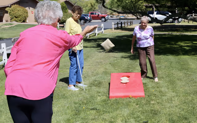 Three elderly women playing cornhole on a grassy lawn outside on a sunny day. One woman in a pink shirt is throwing a bean bag towards a red cornhole board, while another woman in a purple floral shirt watches. A third woman in a yellow shirt and blue pants stands nearby holding a cane.