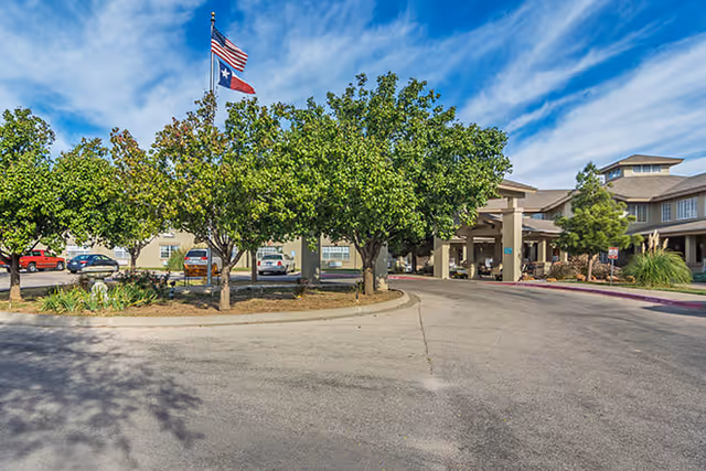 Exterior view of Brookdale Sherwood facility showing the driveway entrance with trees, parked cars, and two flags on a flagpole under a partly cloudy blue sky.