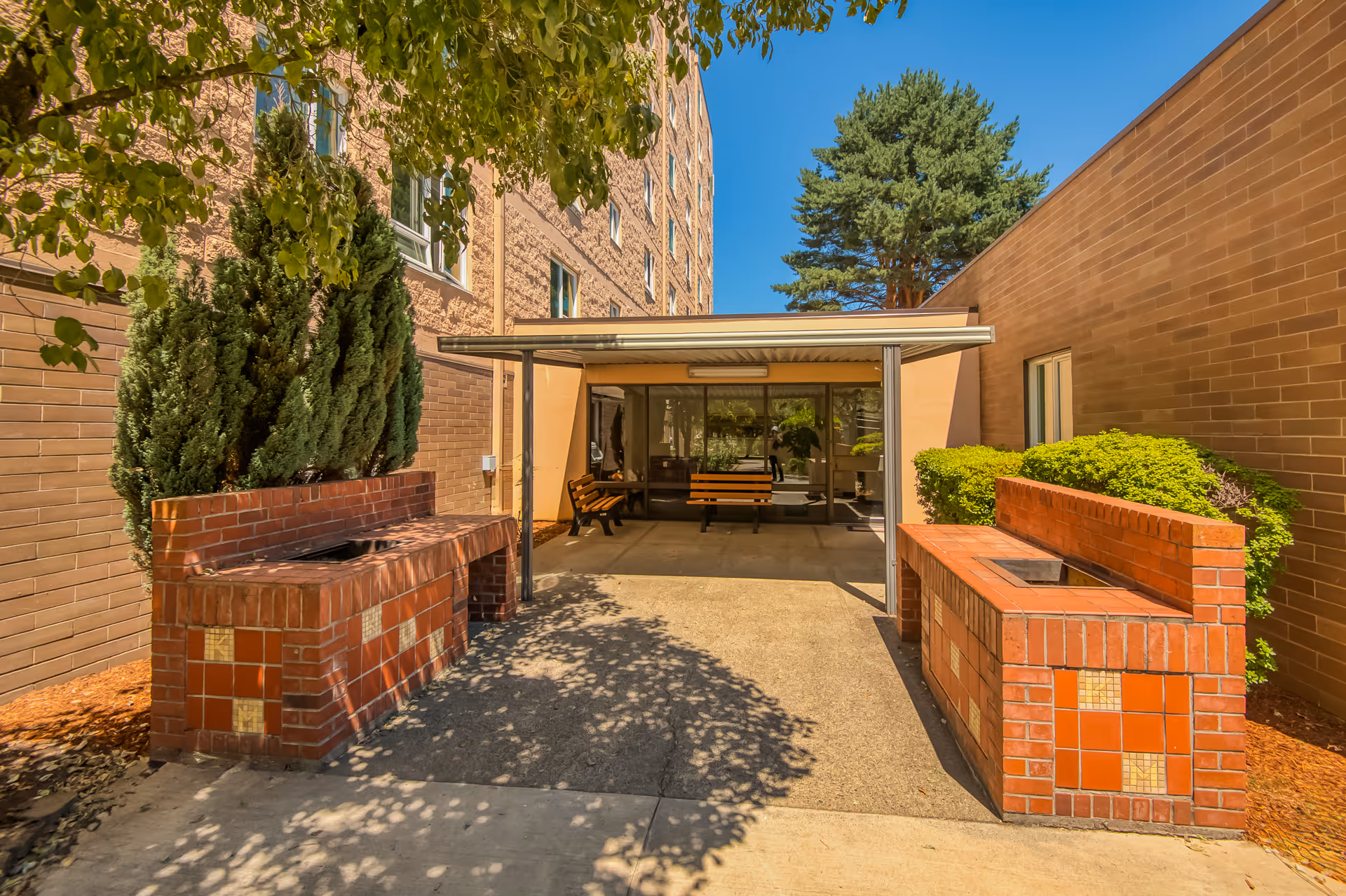 Outdoor patio area at Kirkland Union Manors featuring two brick barbecue grills with tiled accents, surrounded by greenery and trees. The patio is adjacent to a building with large windows and a covered seating area with benches.