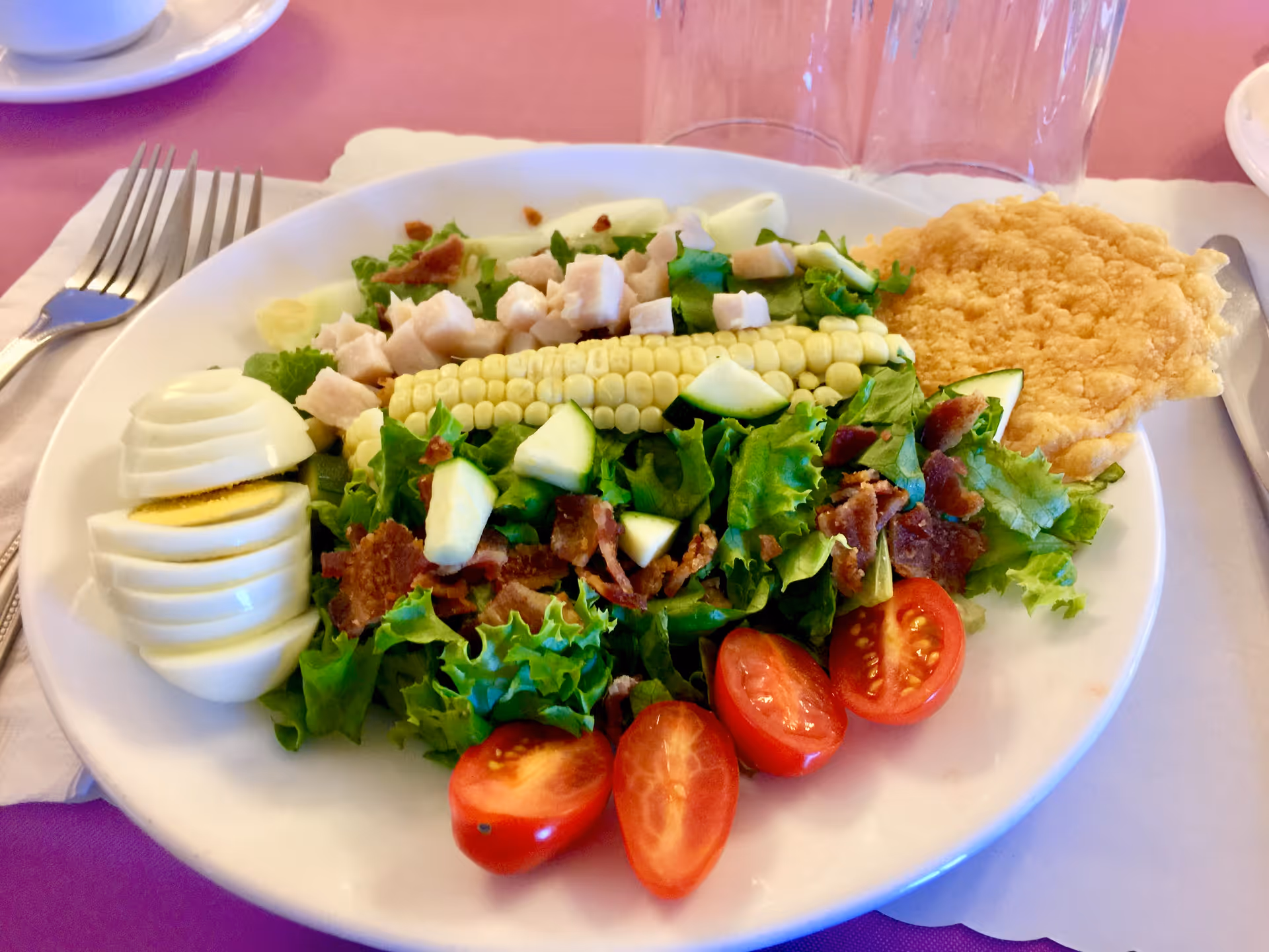 A plate of salad topped with a corn cob, sliced hard-boiled egg, cherry tomatoes, bacon, diced chicken and a cracker on a table.